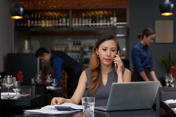 Smiling elegant business lady talking on phone and reading e-mails on laptop screen