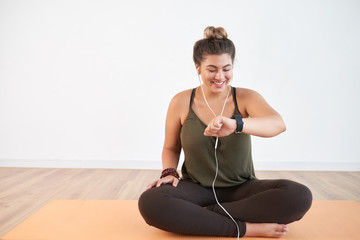 Happy young plus-size woman in earphones sitting on yoga mat and checking fitness tracker