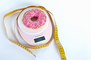 Diet concept. A donut with pink icing lies on a tabletop scale, surrounded by a measuring tape. On a white background.
