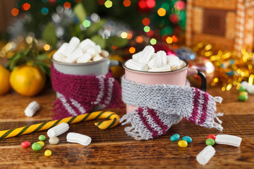 Two mugs in cozy scarves with a hot sweet drink and marshmallows.