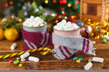 Two mugs in cozy scarves with a hot sweet drink and marshmallows.