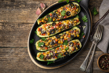 Stuffed zucchini boats with vegetables ( tomato, pepper, corn, red onion and olives) and cheese in cast iron pan, wooden background,  top view