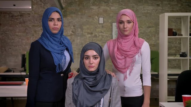 Serious three arab women ar stading and looking straight at camera wearing hihab, middle east working vibes, brick modern office indoor