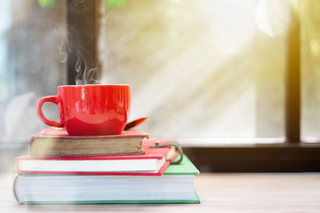 Red coffee cup with smoke on top of stacked old books on wood table with window light. Merry Christmas concept background. Resting, relaxing time or reading.