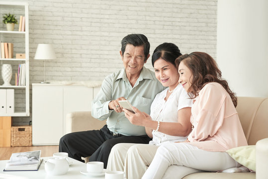 Cheerful aged Vietnamese people resting at home and looking at photos on smartphone