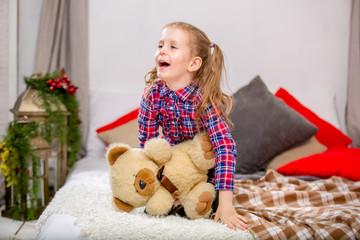 Happy sweet young girl in a checkered blue-red dress sitting on a bed with a teddy bear and laughing