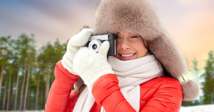 photography, technology and season concept - happy woman in fur hat taking picture by film camera outdoors over winter forest background