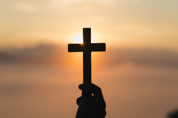 Woman hand holding holy lift of christian cross with light sunset background.