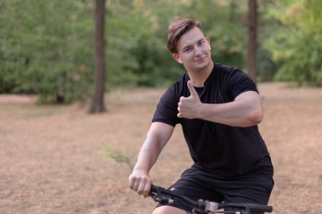 Fototapeta premium Handsome young caucasian man thumbs up biking at the park. White earphones, dark casual wear. Outdoors, abandoned park. Close up, copy space. Smile, emotions and expression. Diversity people.