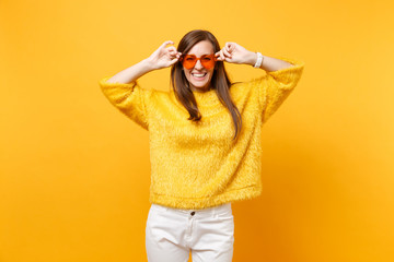 Portrait of laughing happy young woman in fur sweater, white pants holding heart orange eyeglasses isolated on bright yellow background. People sincere emotions, lifestyle concept. Advertising area.