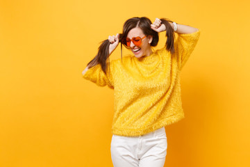 Laughing young woman in fur sweater, white pants, heart orange glasses holding hair like ponytails isolated on bright yellow background. People sincere emotions, lifestyle concept. Advertising area.