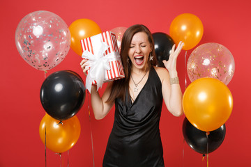 Happy young woman in black dress celebrating screaming, holding red box with gift, present spreading hands on bright red background air balloons. Happy New Year, birthday mockup holiday party concept.