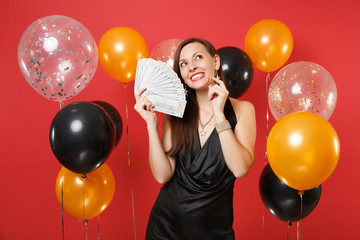 Pretty young girl in black dress holding bundle lots of dollars dreaming about spending cash money on red background air balloons. Valentine's Day Happy New Year birthday mockup holiday party concept.