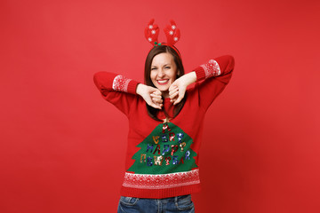 Smiling young Santa girl in knitted sweater fun decorative deer horns on head rising hands isolated on bright red background. Happy New Year 2019 celebration holiday party concept. Mock up copy space.