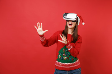 Young Santa girl looking in headset touch something like push click on button, pointing at floating virtual screen isolated on red background. Happy New Year 2019 celebration holiday party concept.