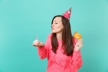 Tender young girl in knitted pink sweater, birthday hat with closed eyes blowing out candle on cake, hold in hand credit card isolated on blue background. People lifestyle concept. Mock up copy space.