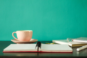 Desk with an open spiral notebook, pen pile of notebooks, and coffee: front view. Blue background, free copy space, selective focus