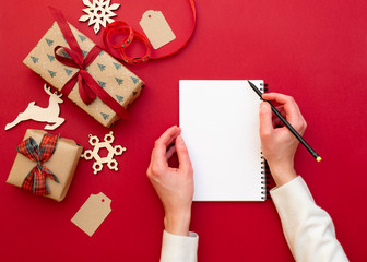 Women's hands holding pencil and notebook with wish list on red background with Christmas decorations and gifts. Xmas and Happy New Year composition. Flat lay style, top view, overhead, copy space