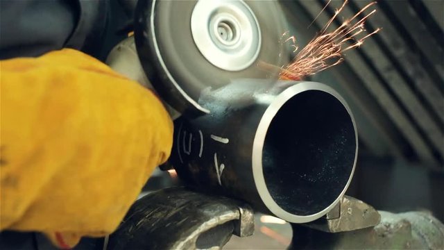 A yello-gloved mechanic cleans a welded seam on a section of a steel pipe with the help of a grinding machine in the metal workshop