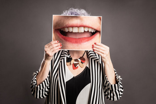 Close up portrait of beautiful older woman holding a picture of a mouth smiling on a gray background