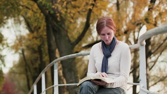A young woman reads the Bible and prays in a park by autumn.