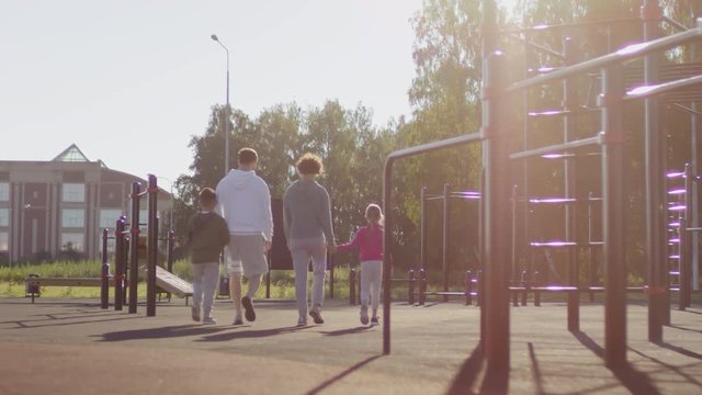 Rear view of mother and father holding hands with little daughter and son while walking together on outdoor workout playground at summer day