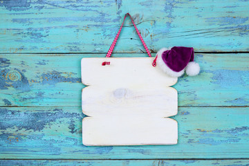 Blank white wood sign hanging by ribbon on antique rustic teal blue wood door with purple Christmas Santa Claus hat; holiday background with painted copy space