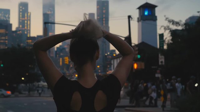 TRACKING Attractive mid-30s Caucasian female braids her hair before jogging in the evening, Downtown NYC Manhattan in the background. 4K UHD