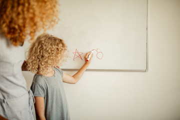 Schoolgirl in home wear writing something on white board on the wall with a marker and her mother...