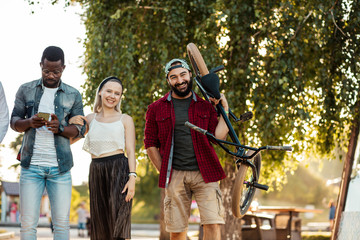 Young BMX rider in company with his multiethnic friends walking to have a good time at the city park