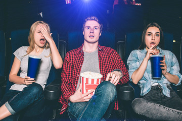 Naklejka premium Youn man and women are sitting in chairs and watching movie. They are surprised and confused. Blonde girl put her hand on face. Brunette girl stopped drinking cola. Guy is holding a basket of popcorn.