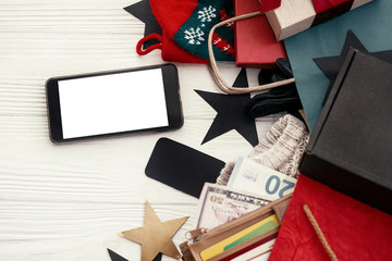 Christmas shopping and seasonal sale. Credit cards and money in wallet, phone with empty screen, bags, clothes, gift boxes, tags on rustic wood. Space for text. Advertising app