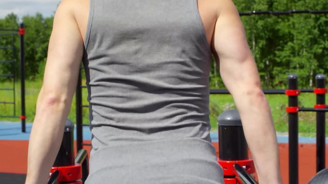Rear view of muscular man in sportswear doing dips on parallel bars while training on outdoor workout playground on summer day