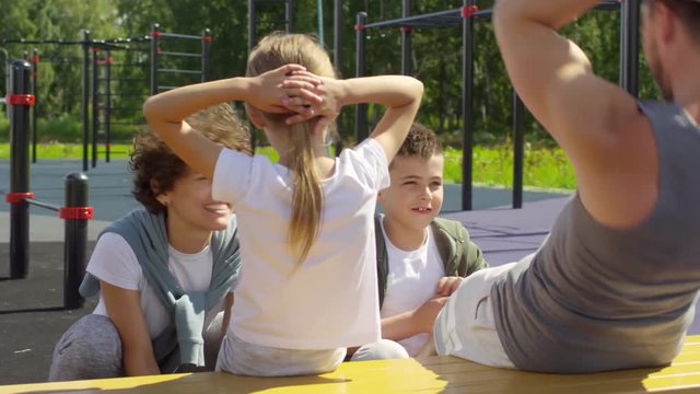 Happy family having outdoor workout on playground in the park: cheerful mother and little boy holding legs of father and daughter and supporting them while doing sit ups on bench
