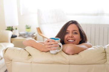 Perfect smile Beautiful and young brunette girl laughing and happy taking a cup of coffee or tea while stay on the sofa of her house in the bright living room
