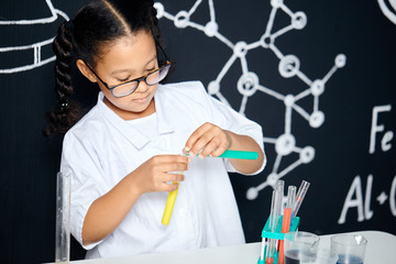 Mixed race asian, african girl in protective glasses wearing white lab holding test tubes and flasks conducting experiments over black board with symbols.