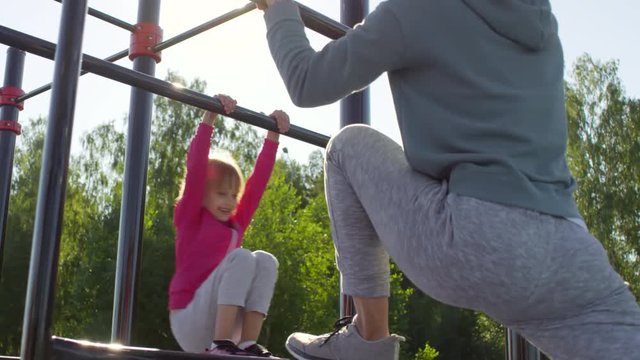 Sporty mother doing stretching exercise while little daughter smiling and playing on jungle gym outdoor on sunny summer day