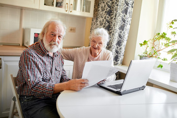 Mature couple sitting at kitchen table with laptop looking through financial papers, having little jam with the pension contributions.
