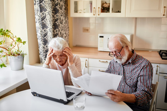 Mature Couple Sitting At Kitchen Table With Laptop Looking Through Financial Papers, Having Little Jam With The Pension Contributions.