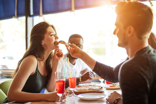Young multiracial diverse colleagues eating pizza during their lunch time at office pizzeria, playing team building game, feeding his partner with tasty pizza