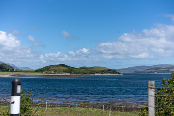 Sicht von der Straße zwischen zwei Pfosten sieht man einen Zaun Weide und dahinter ein See,  ein paar weißen Wolken am blauen Himmel Schottland