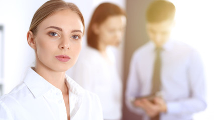 Fototapeta premium Beautiful blonde businesswoman standing straight in a brightly lit office at the background of colleagues or partners. Business concept
