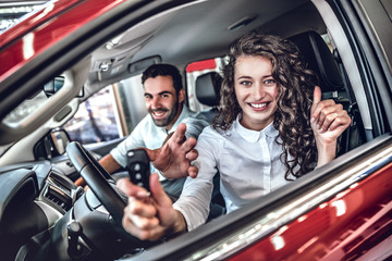 Close up of a happy young couple smiling showing car keys and thumbs up in a new car at the dealership