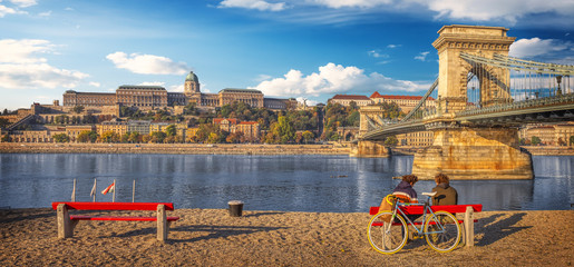 A couple enjoying the view while sitting on a bench near the Danube river embankment in Budapest,...