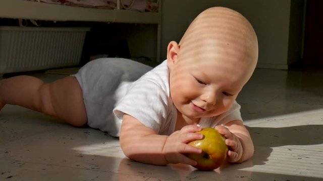Baby is lying on the floor and holding a red apple. The child is very lively and cheerful, he actively knocks with hands and feet on the floor