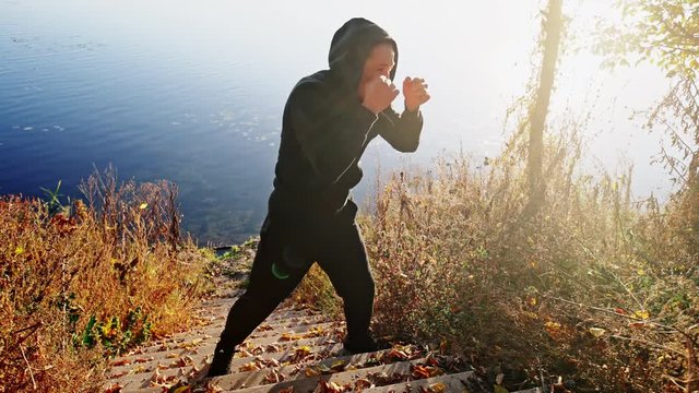 Young active man trains in a box against the background of a lake in a cool autumn morning. Boxing training outdoor.