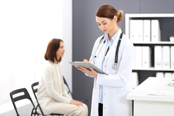 Doctor woman at work. Portrait of female physician filling up medical form while standing near reception desk at clinic or emergency hospital. Patient woman sitting at the background. Medicine concept