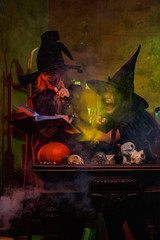 Photo of laughing two witches in black hats reading book of incantations on cauldron with steam