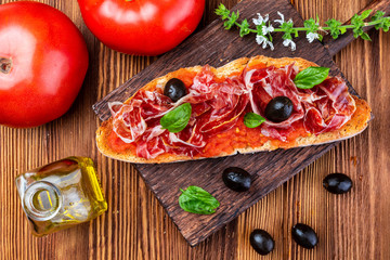 Delicious bread toast with natural tomato, extra virgin olive oil, Iberian ham, black olives and basil leaves. On wooden background.