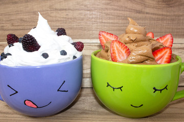 Two large mugs with painted faces, whipped cream and berries stand on a wooden shelf. Chocolate mousse with strawberries and vanilla mousse with blackberries and blueberries.